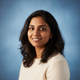 Portrait of a smiling Indian woman, late 20s, against a soft sky blue background.
